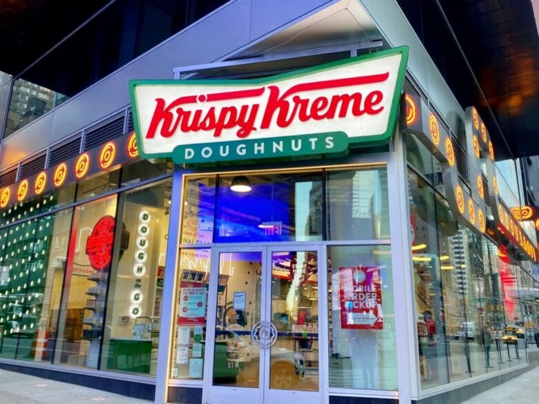 Fachada iluminada de una tienda Krispy Kreme Doughnuts en Nueva York, con el logotipo de la marca sobre la entrada.