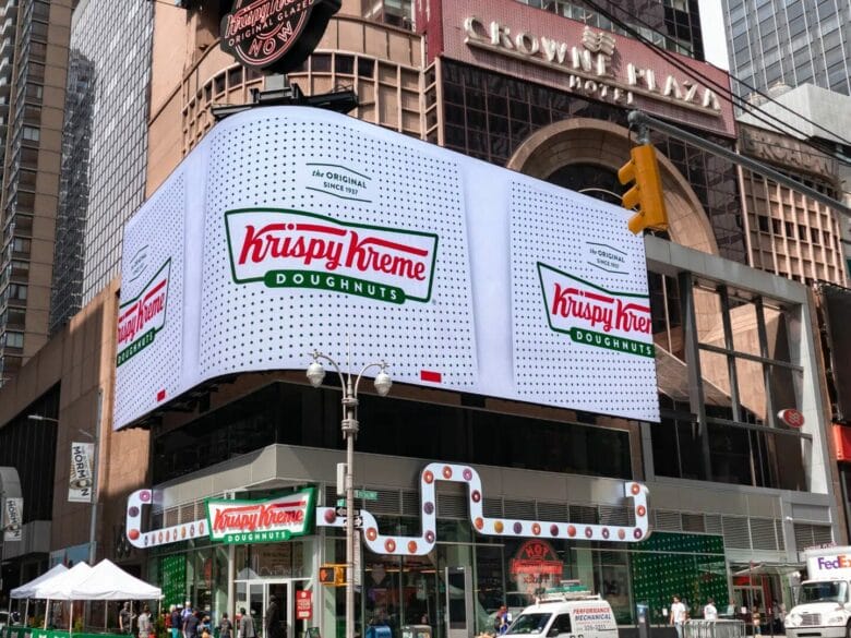 Fachada de Krispy Kreme en Times Square, Nueva York, con pantallas gigantes, el logotipo de la marca y la entrada de la tienda en plena ciudad.