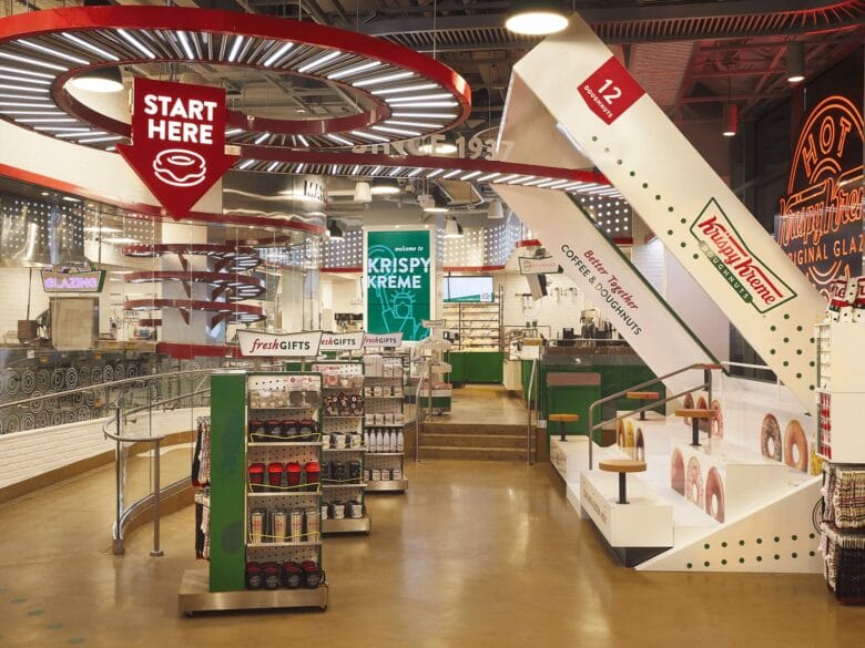 Interior de una tienda Krispy Kreme en Nueva York con zona de pedidos, decoración temática, cartel de Start Here y una gran caja de dónuts.